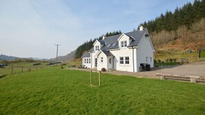 Une grande maison blanche avec un toit gris, située sur une colline herbeuse, avec une forêt en arrière-plan.