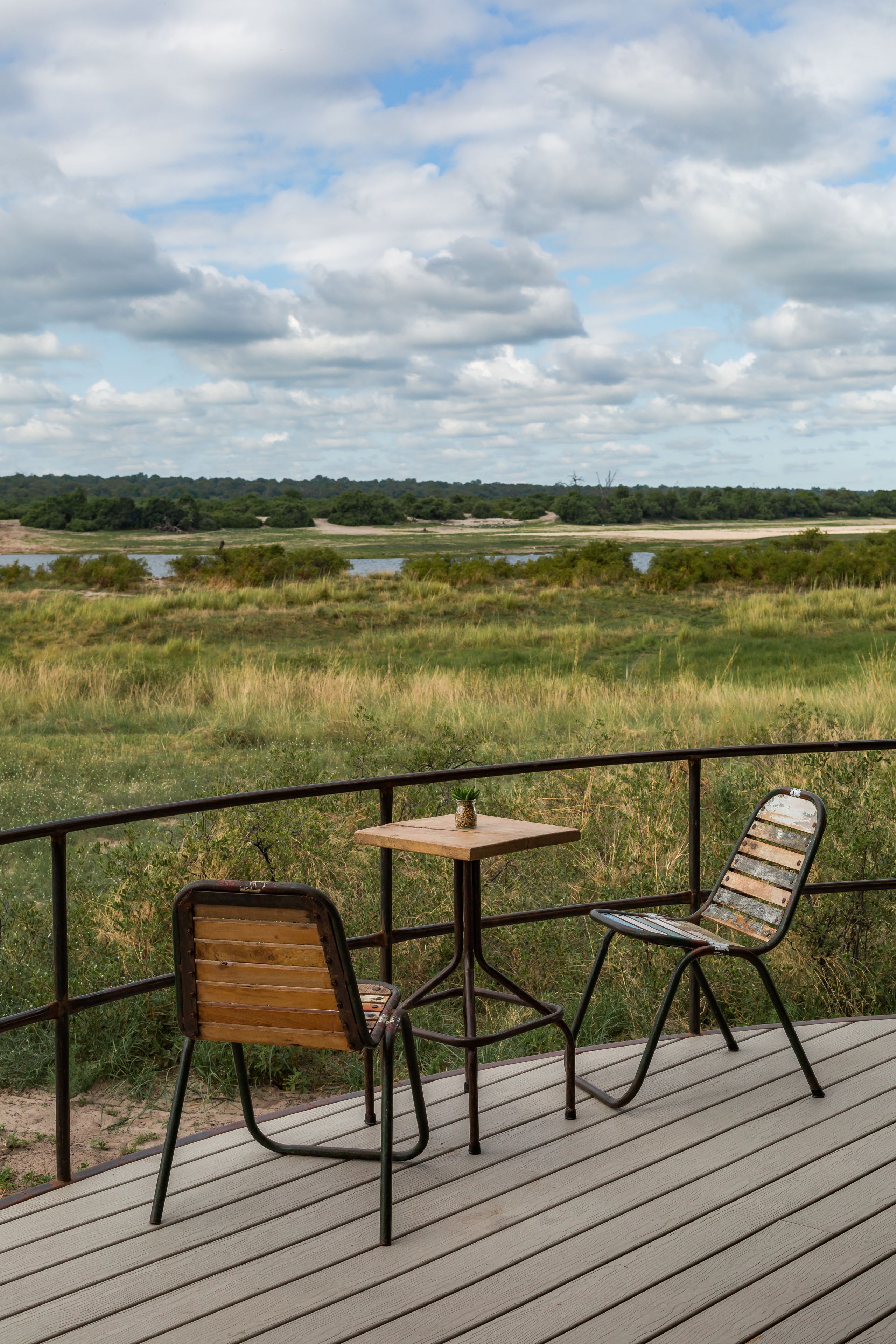 A deck with two chairs and a table overlooking a grassy landscape with a water body and trees in the distance.