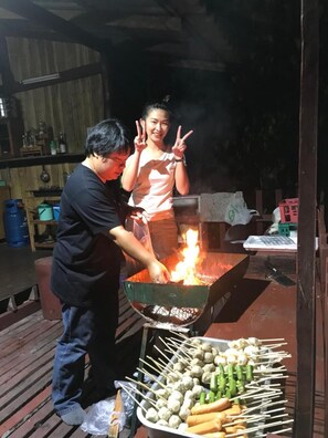 A person grilling food outdoors with a fire visible in a grill. - Forest Guesthouse (Samoeng)