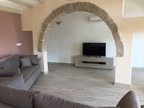 A living room with a stone arch, a grey sofa, a flat-screen TV on a white stand, and a grey tiled floor. - Beautiful villa with amazing swimming-pool, seafront (Okrug Gornji)