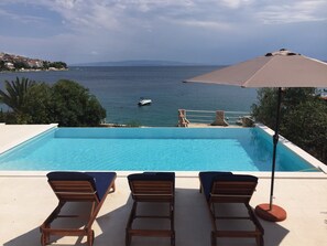 A rectangular swimming pool with three wooden lounge chairs and a large umbrella.