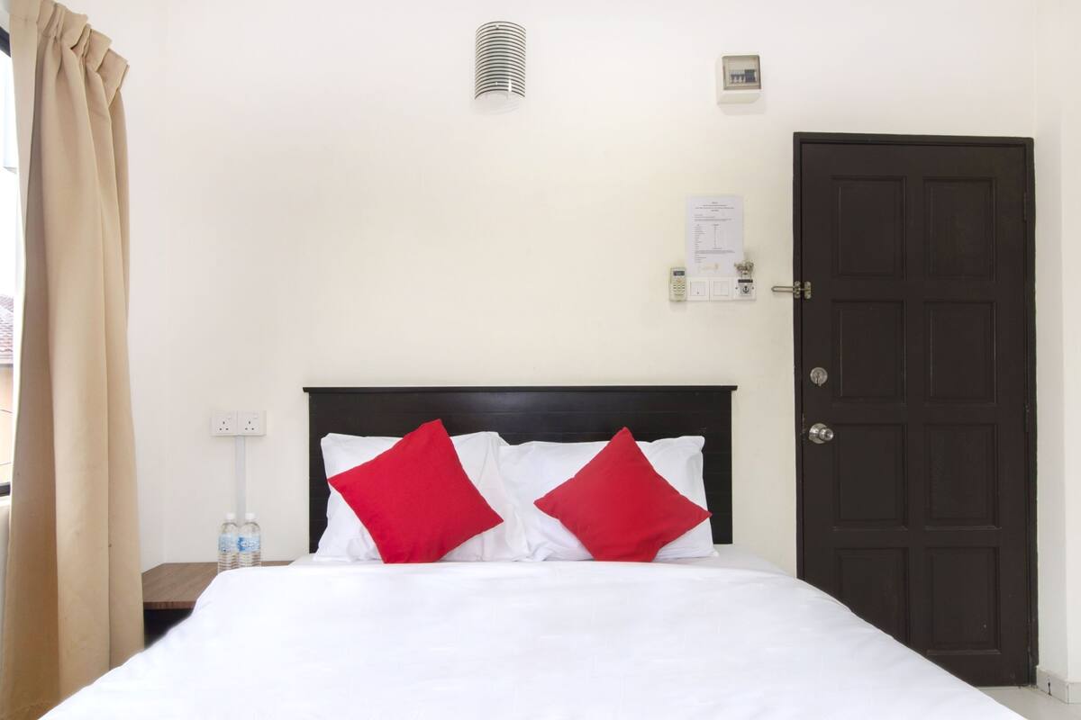 A hotel room with a bed, a dark wooden door, a light switch, and a power outlet on the wall.
