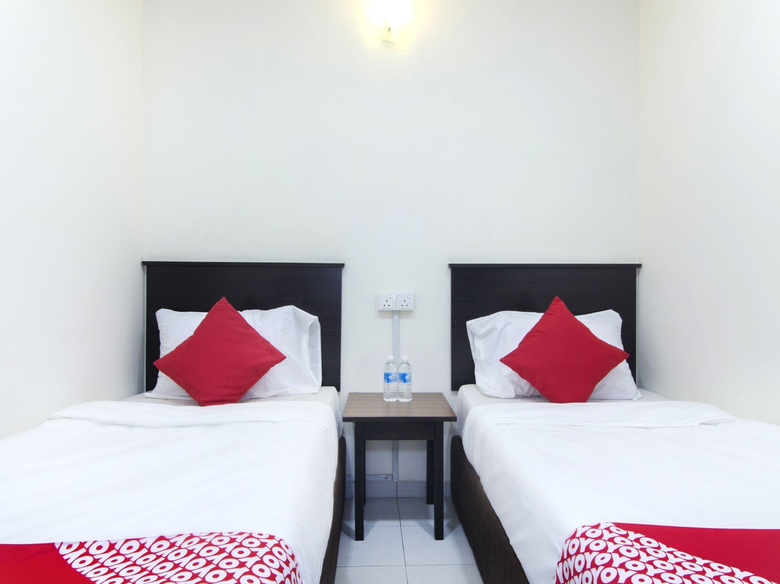 A hotel room with two beds, each with a black headboard, white bedding, and red pillows. There is a small wooden nightstand between the beds with a water bottle and glasses on it.