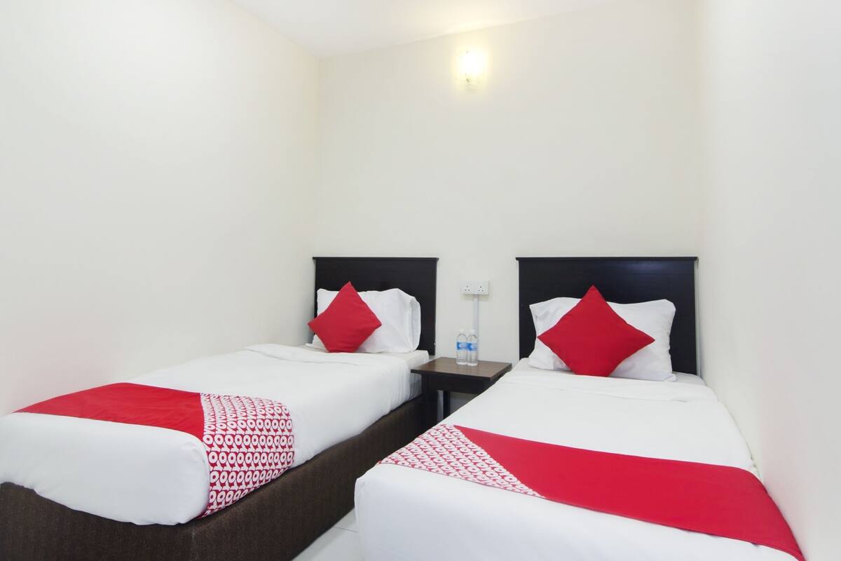 A hotel room with two single beds, each with a red and white patterned bedspread and matching pillows. There is a small table between the beds with a water bottle on it.