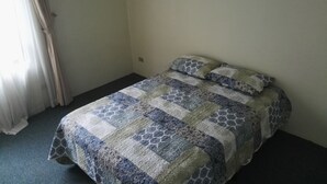 Un lit simple avec une couvre-lit à motifs et deux oreillers, une fenêtre avec des rideaux et deux tableaux encadrés accrochés au mur.