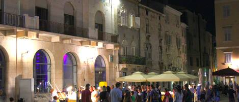 A crowded outdoor square with historic buildings, outdoor seating, and umbrellas.
