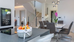 Een moderne keuken met een centraal eiland, een eethoek met een witte tafel en donkere stoelen, en een trap met een glazen balustrade.
