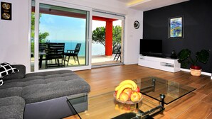 A modern living room with a sofa, a glass coffee table with fruit, a TV, and a view of the sea through large glass doors. - Villa With Private Pool (Kukljica)