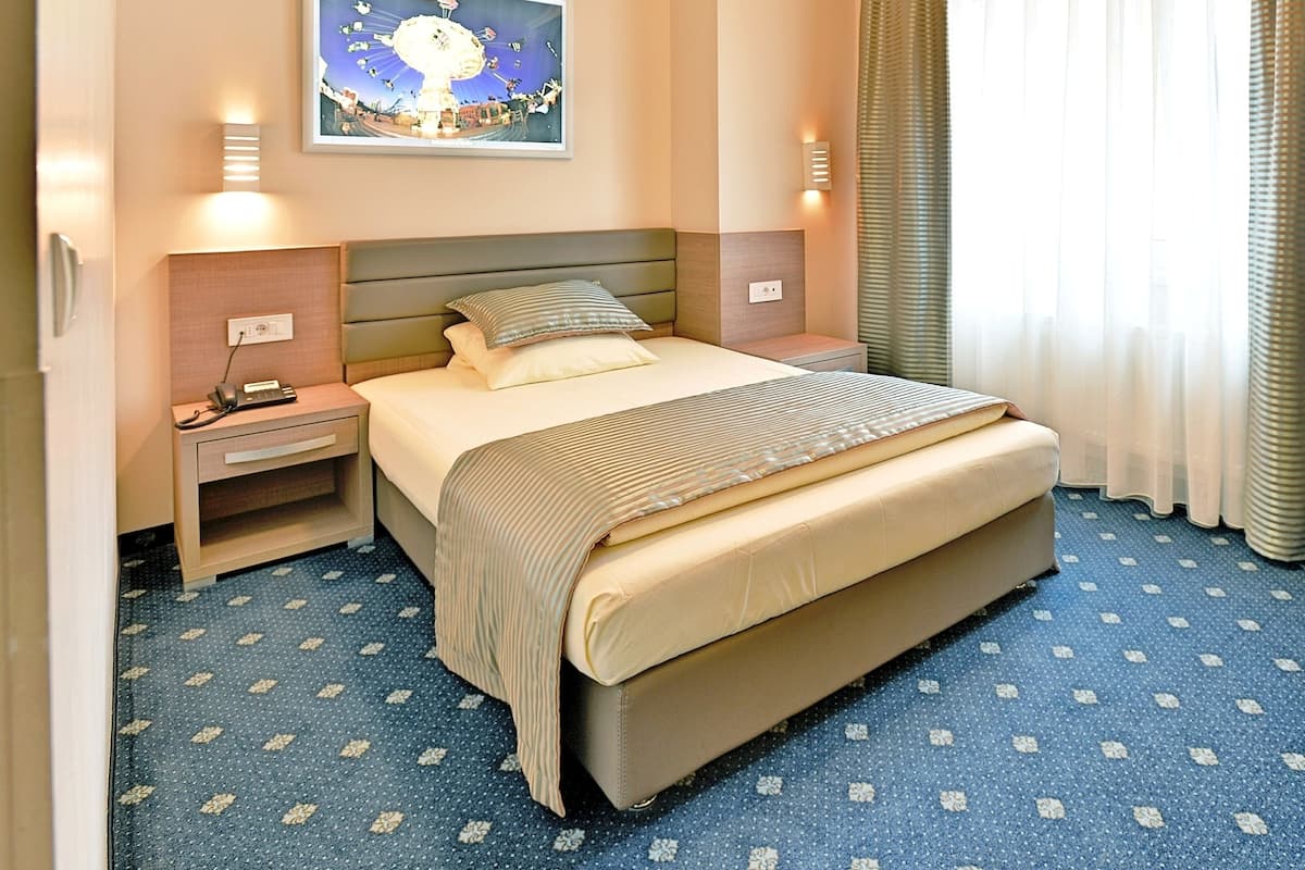 Ein Hotelzimmer mit einem Bett, einem Nachttisch, einem Telefon, einem Bild an der Wand und einem Fenster mit Vorhängen.