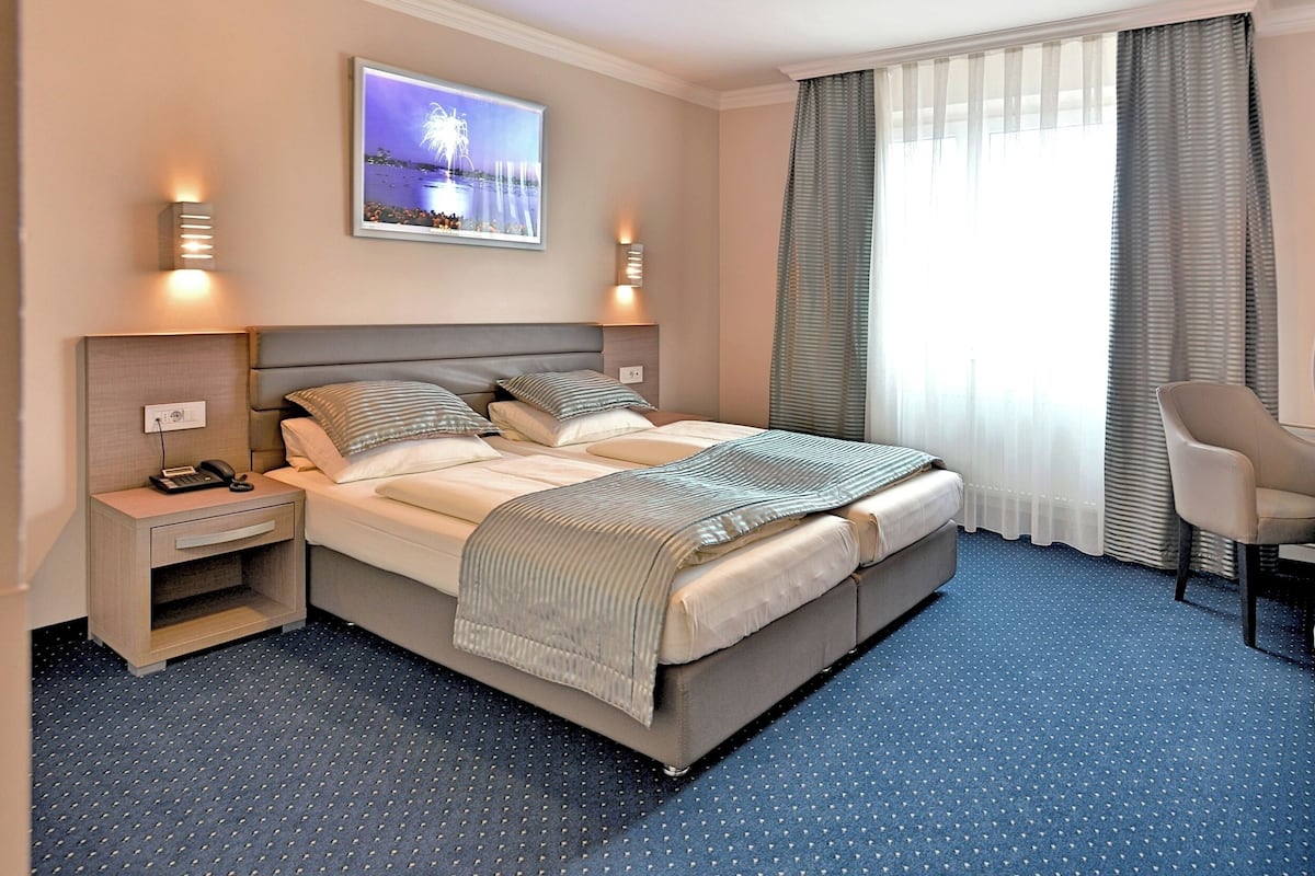 Ein Hotelzimmer mit einem großen Bett, einem Schreibtisch mit Telefon, einem Sessel und einem Fenster mit Vorhängen.