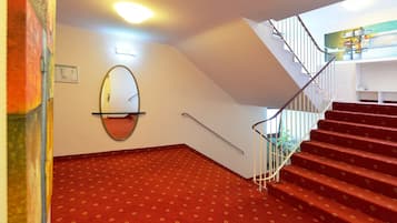 Un escalier avec des marches recouvertes de moquette rouge et une rampe en métal, menant à un palier agrémenté d’un miroir mural et d’un luminaire.