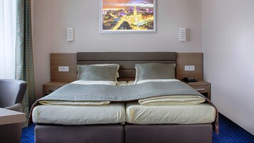 A double bed with striped bedding, two bedside tables, a chair, and a cityscape painting on the wall.