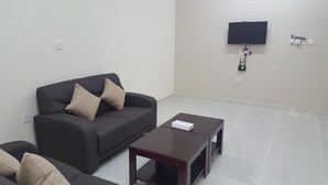 A living room with a dark sofa, two beige cushions, a small wooden coffee table, and a flat-screen TV mounted on the wall. - Today House Suites (Salalah)