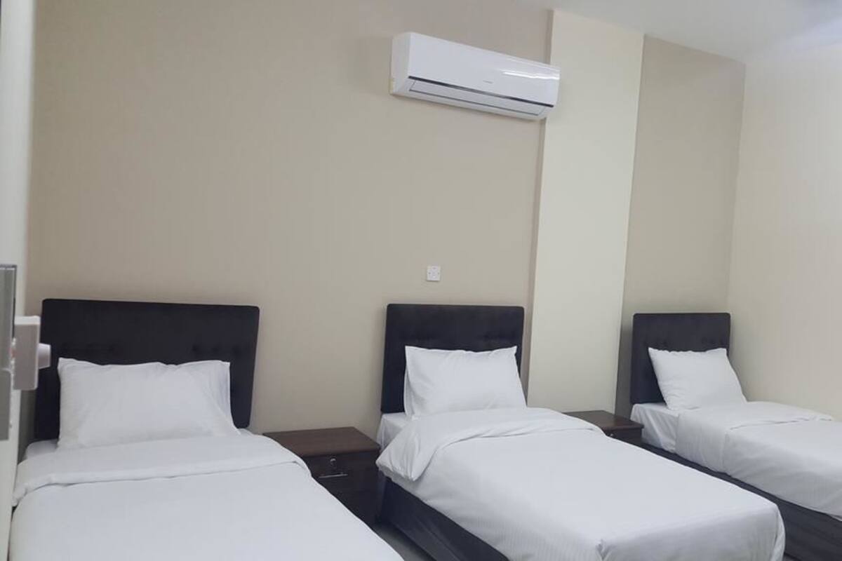 A hotel room with three single beds, each with a dark headboard and white bedding. An air conditioning unit is mounted on the wall above the beds.