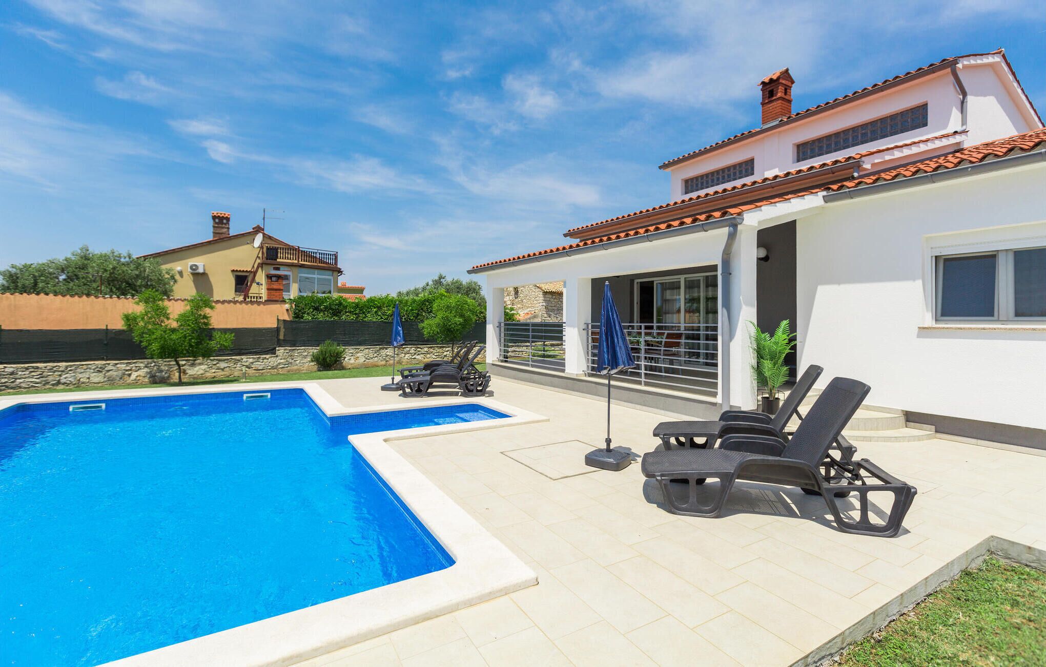 Une piscine avec des chaises longues et des parasols, un bâtiment blanc avec un toit en tuiles et un mur de pierre en arrière-plan.
