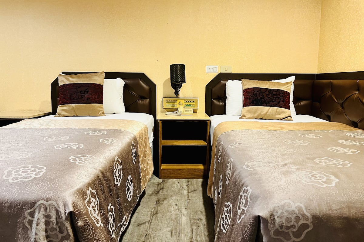 A hotel room with two beds, each with a patterned bedspread and pillows. There is a bedside table with a lamp and a radio.