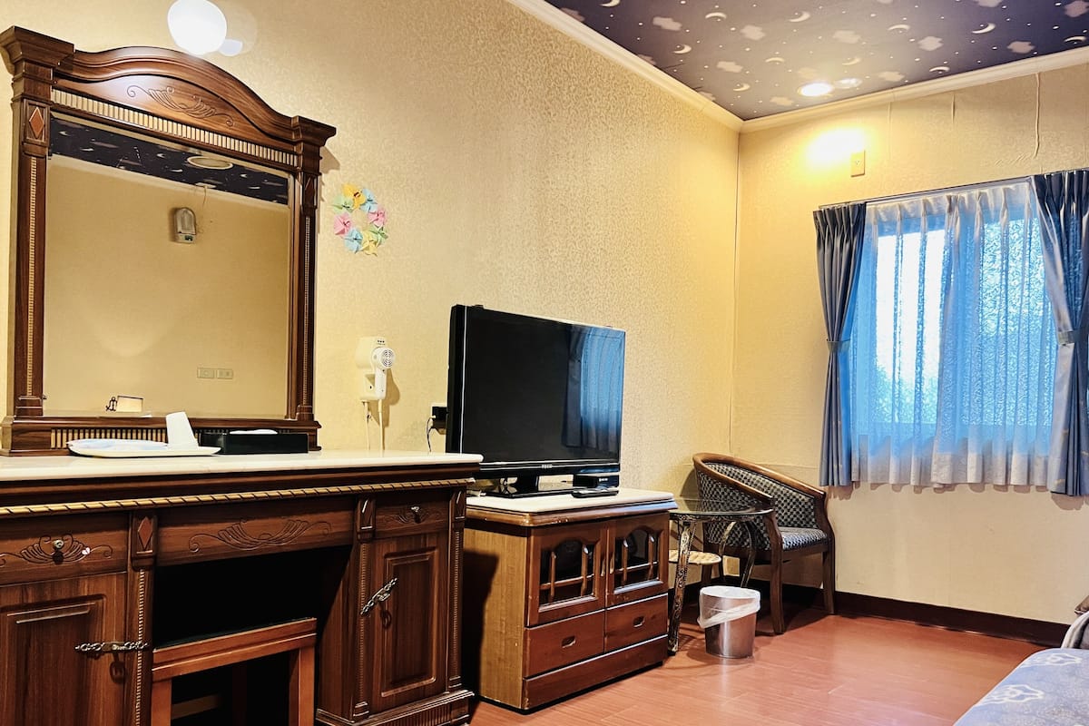 A hotel room with a wooden dresser, a flat-screen TV, a mirror, a window with curtains, and a bed.