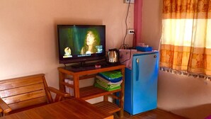 A room with a wooden table, a blue refrigerator, a television displaying a program, and a bed with a purple blanket. - Wongwan Farm (Samoeng)