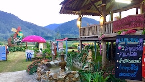 A rustic outdoor seating area with stone walls, potted plants, and a windmill in the background. - Wongwan Farm (Samoeng)