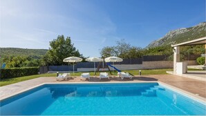 Una piscina rectangular con agua azul cristalina, rodeada de sillas de descanso blancas y sombrillas. Hay escalones para entrar a la piscina y un tobogán de madera. La piscina tiene como fondo colinas verdes y un cielo despejado.