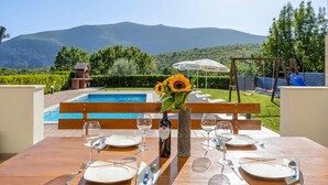 Mesa de comedor para cuatro personas con vista a una piscina y montañas al fondo.