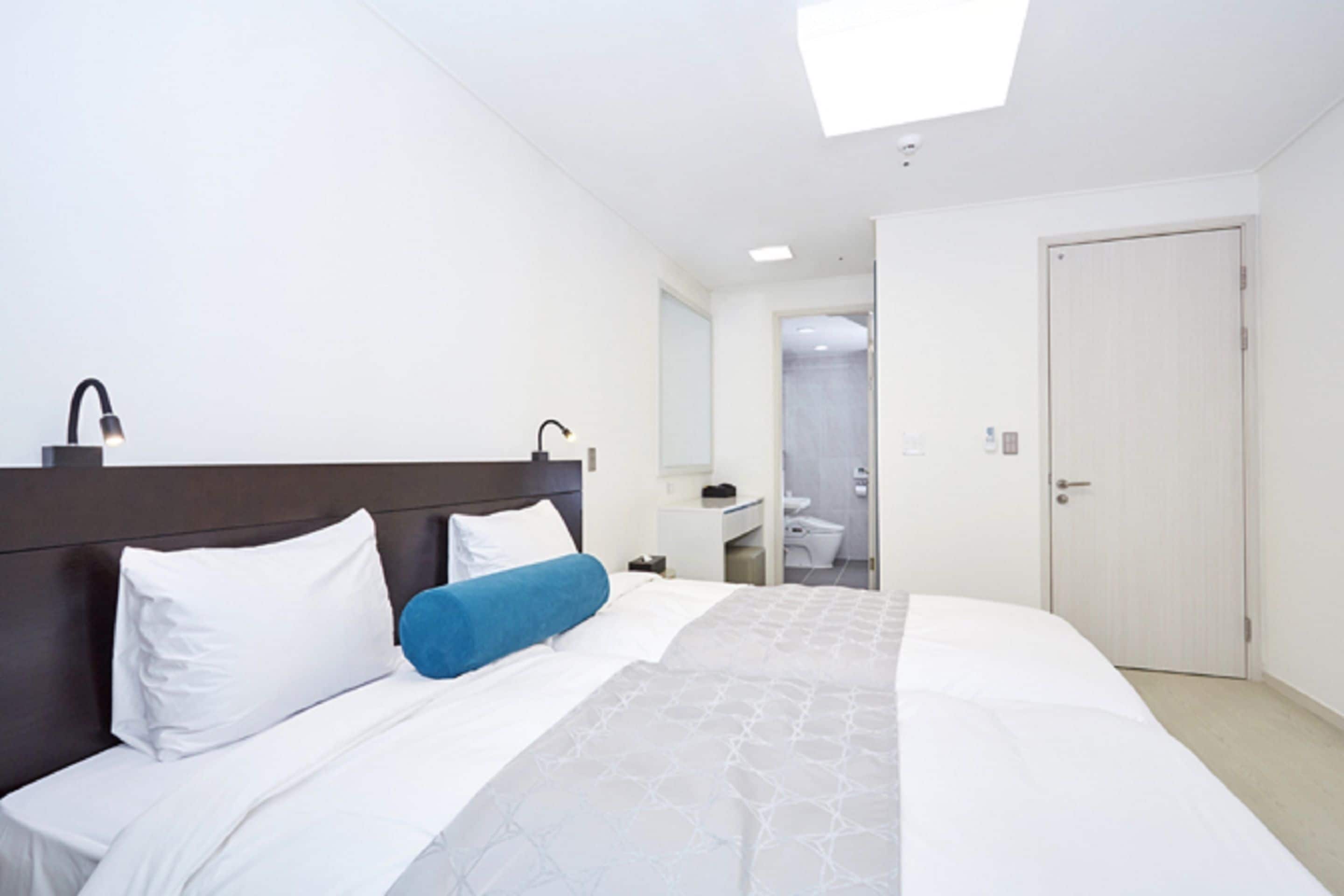 Ein Hotelzimmer mit Bett, Nachttisch, Spiegel, einem durchgegangenen Tür erkennbaren Badezimmer und einer Deckenleuchte.