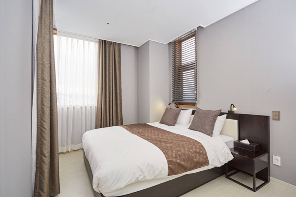 A modern hotel room with a large bed, a nightstand, and a window with blinds.