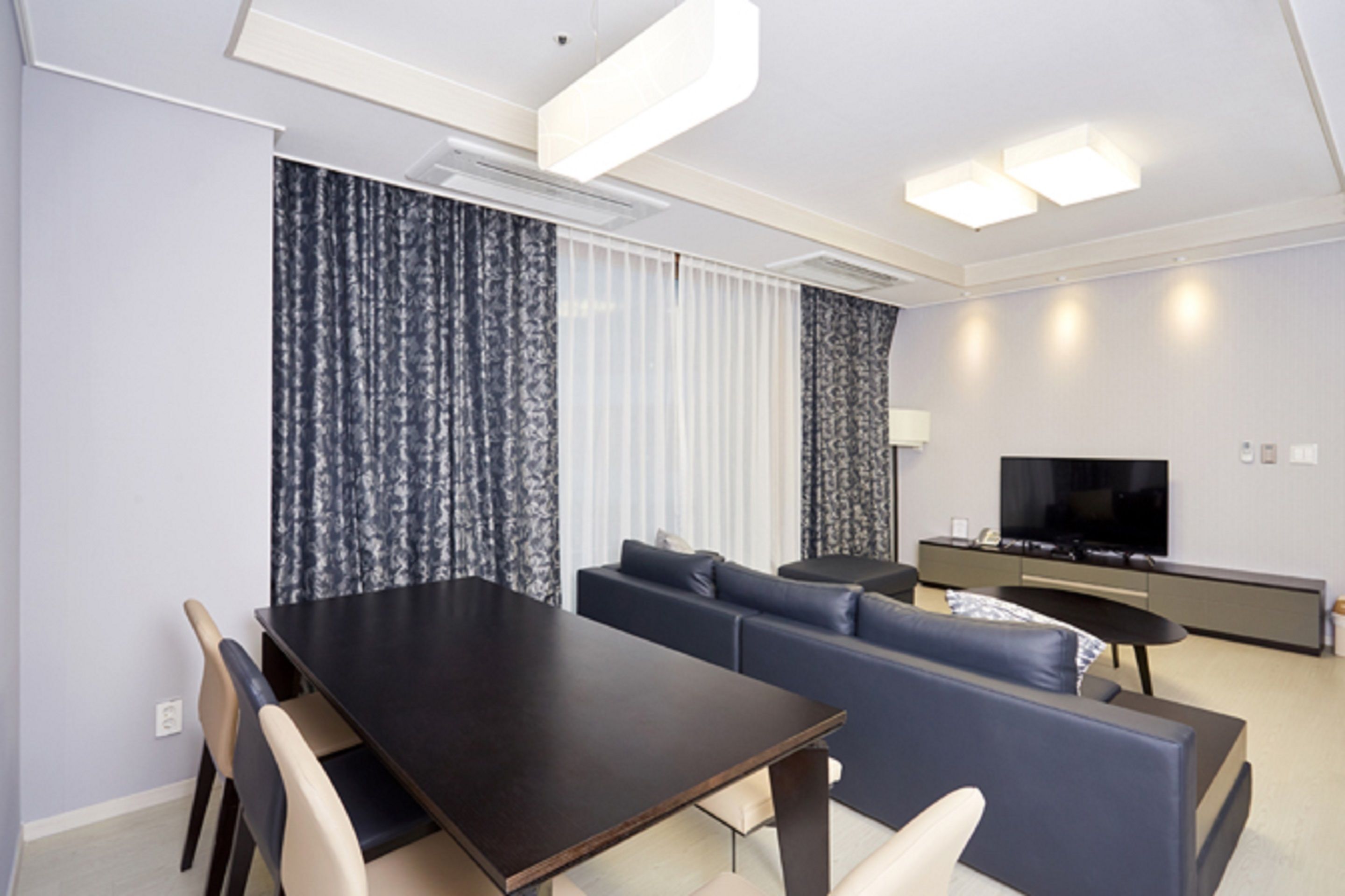 A modern living room with a dark wooden dining table, beige chairs, a blue sofa, and a flat-screen TV on a grey console.