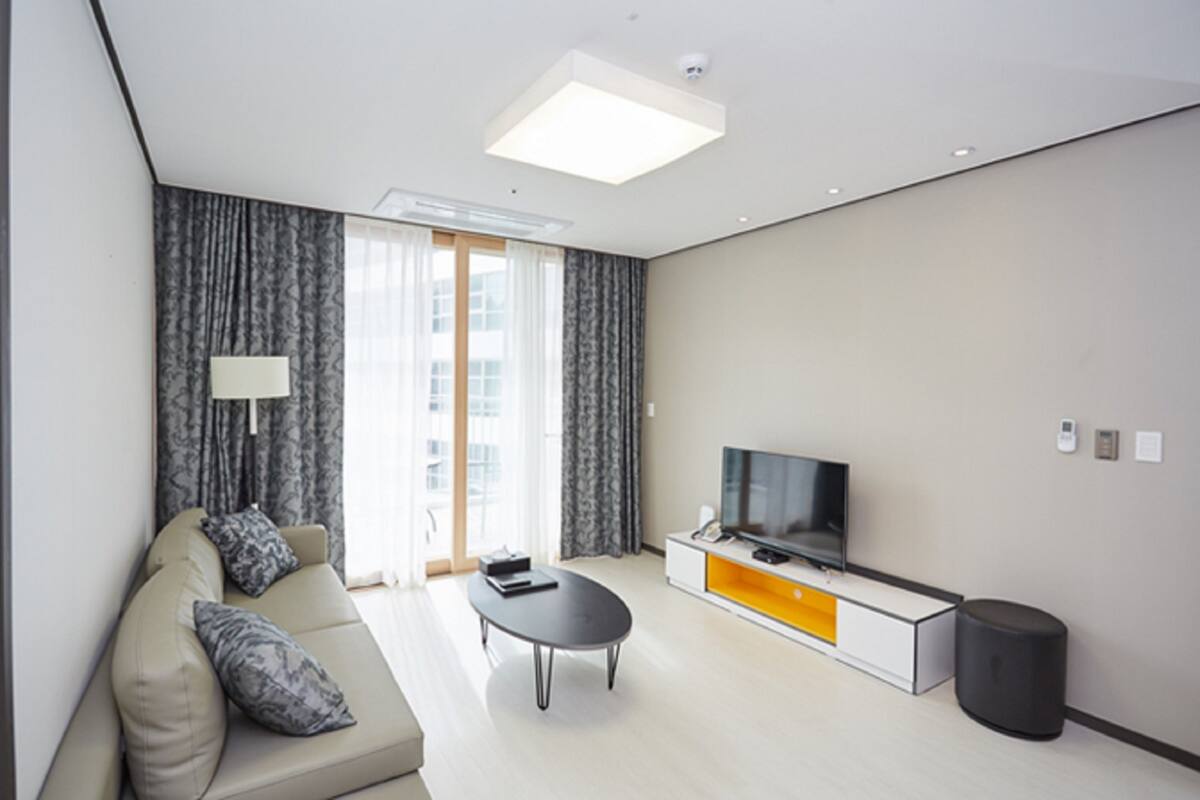 A modern hotel room with a sofa, a coffee table, a TV on a white cabinet, and a large window with curtains.
