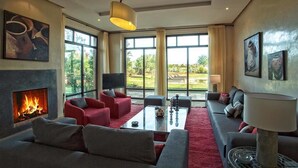 A modern living room with a fireplace, red and grey sofas, a coffee table, and large windows offering a view of the outdoors. - Villa De Luxe Samanah (Marrakech)
