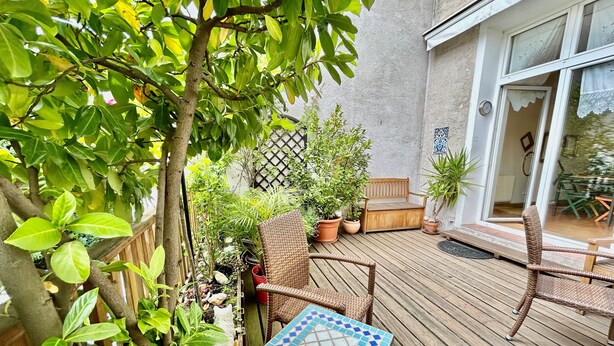 Une terrasse en bois, agrémentée de chaises en osier et d’une table à motifs de mosaïque, entourée de plantes en pot et d’une clôture en bois.
