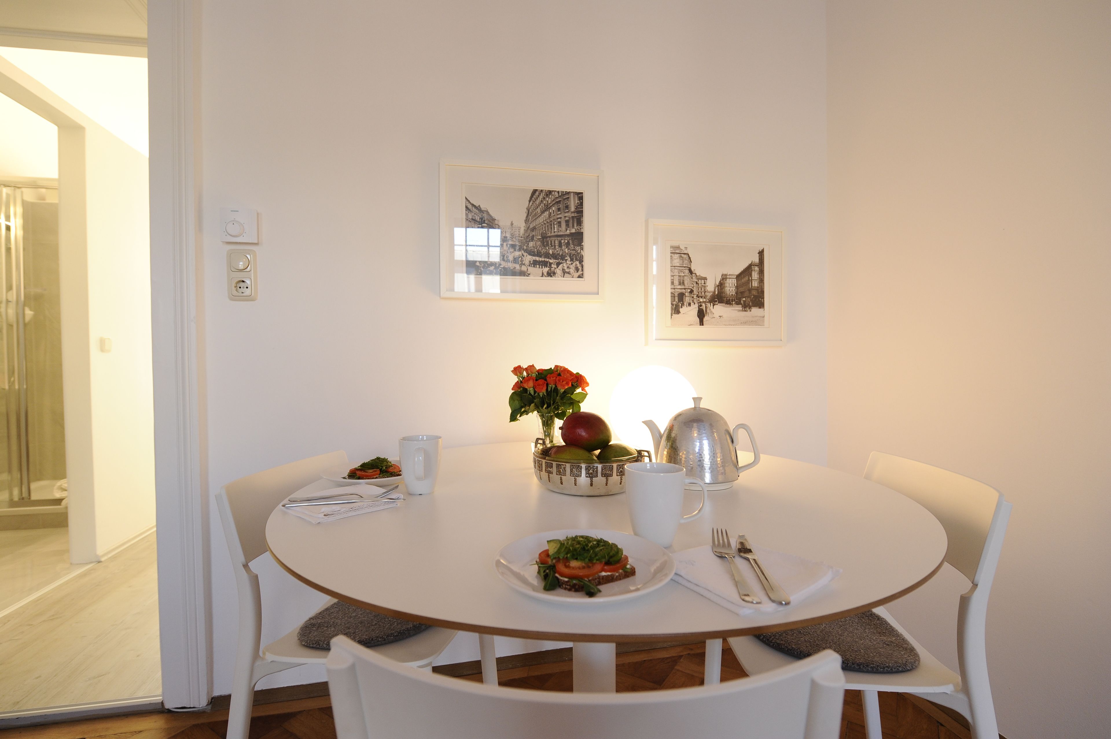 Une table de salle à manger ronde, entourée de chaises blanches, avec un pichet blanc, un saladier de fruits et une assiette de nourriture. Deux tableaux encadrés sont accrochés au mur, au-dessus de la table.