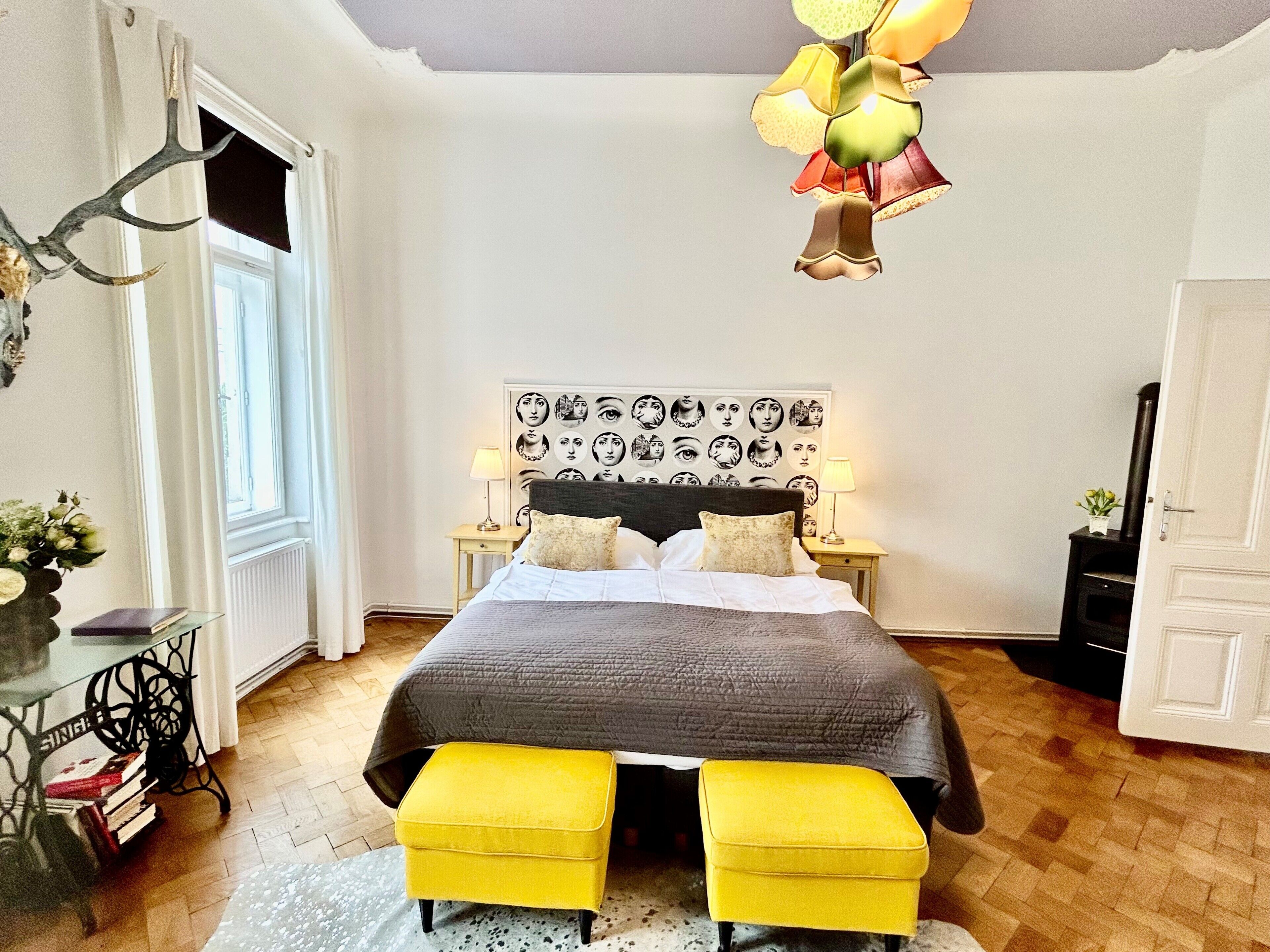 Ein Schlafzimmer mit einem Bett, zwei gelben Hocker, einem Wandbild, einer Lampe und einem Kamin.