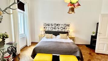 Ein Schlafzimmer mit einem Bett, zwei gelben Hocker, einem Wandbild, einer Lampe und einem Kamin.