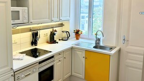 Une cuisine compacte avec des meubles blancs, un meuble inférieur jaune, un four encastré, un four à micro-ondes, une bouilloire et un évier surmonté d’une fenêtre.