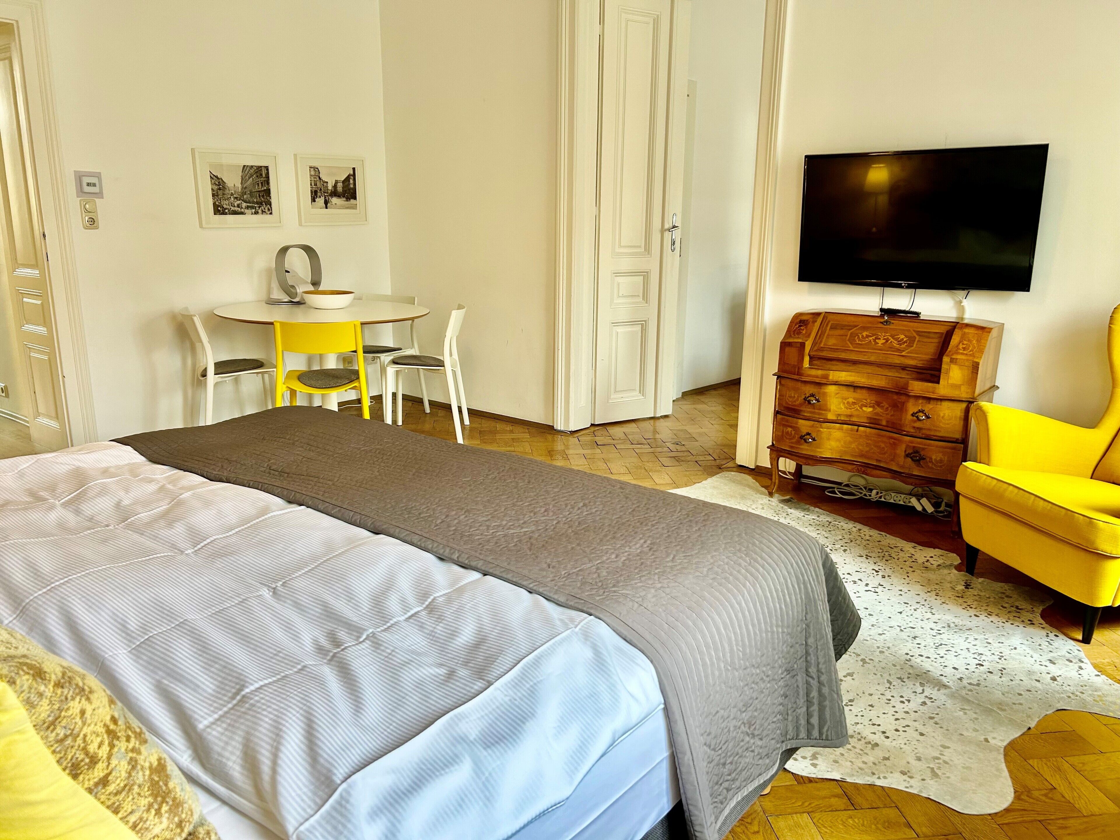 A hotel room with a bed, a TV, a wooden dresser, a yellow chair, a small table with chairs, and two framed pictures on the wall.