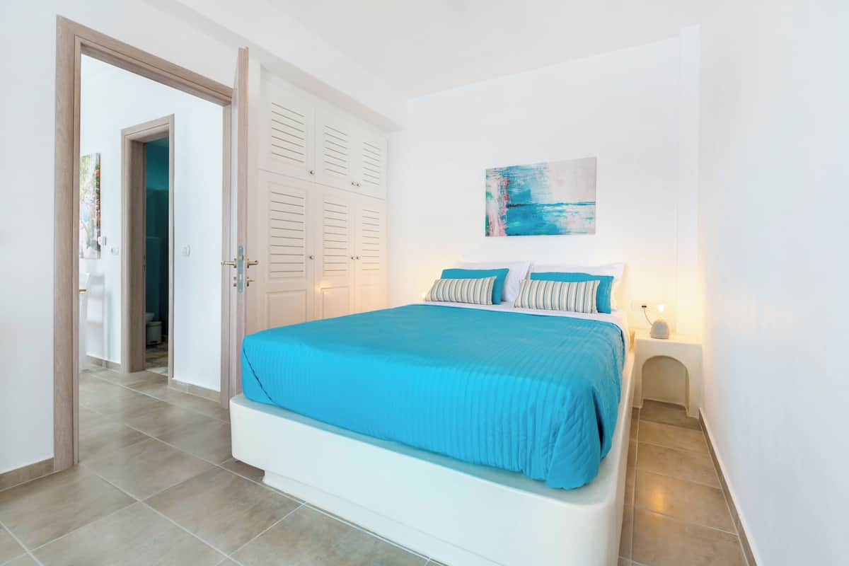 A bedroom with a bed, a nightstand, a painting, and a door leading to another room.