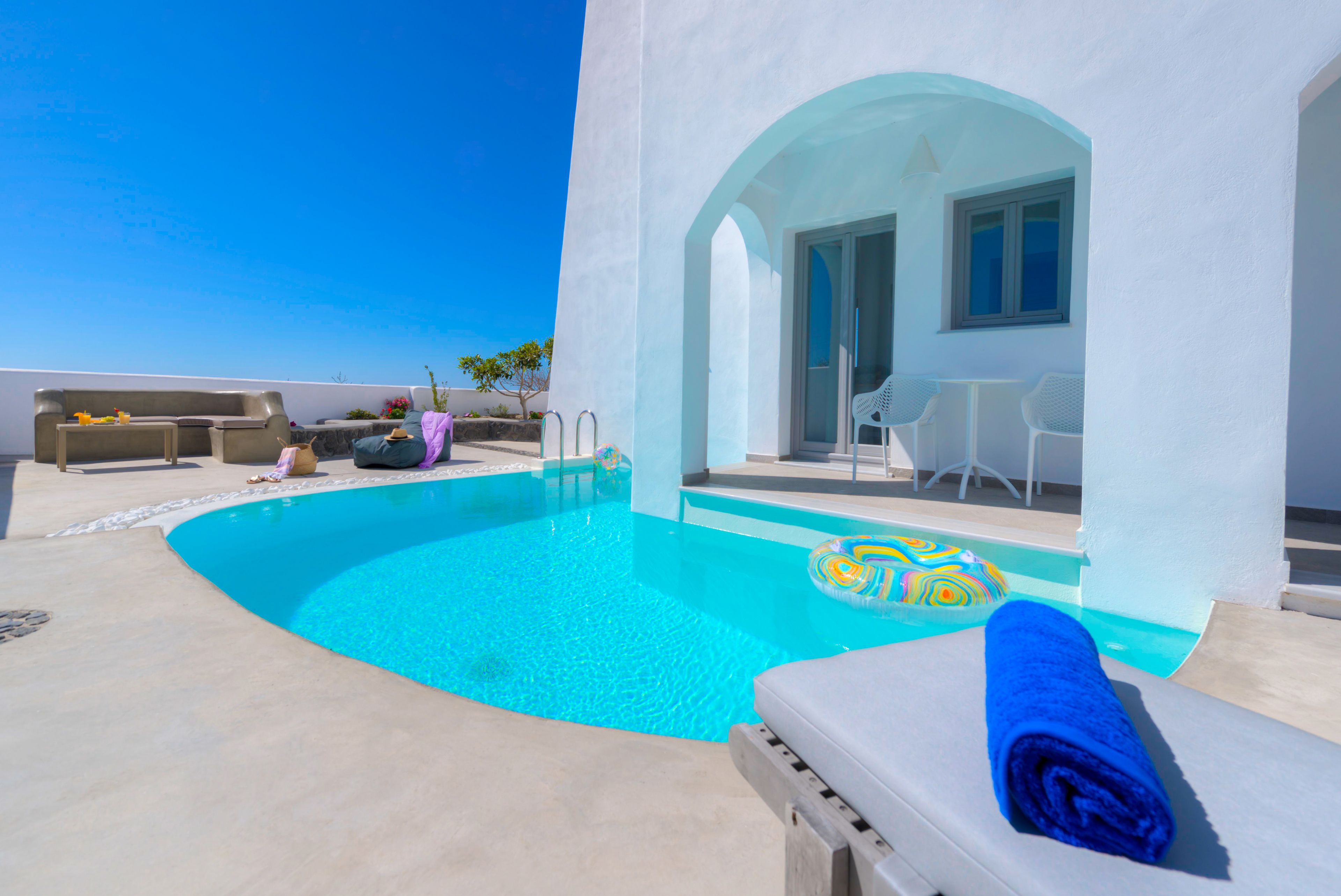 A pool area with a clear blue pool, a lounge chair with a blue towel, and a white building with a door and windows.
