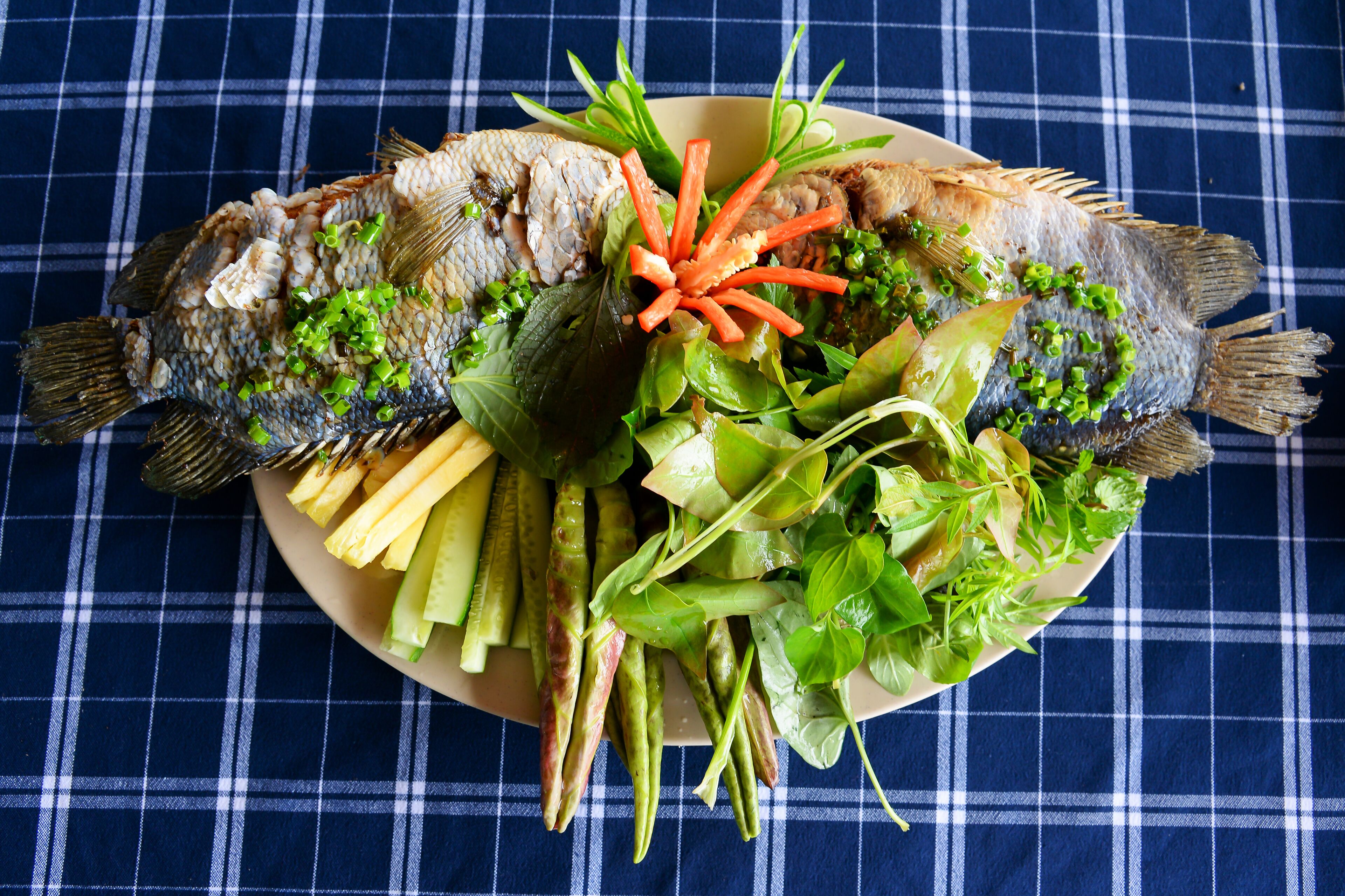 Une assiette contenant deux poissons entiers, décorée de ciboulette, de carottes et de légumes verts à feuilles.