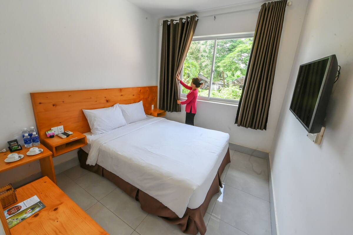 Ein Hotelzimmer mit einem großen Bett, einem hölzernen Kopfteil, Nachttischen mit Annehmlichkeiten, einem fenster mit Vorhängen und einem Fernseher an der Wand.