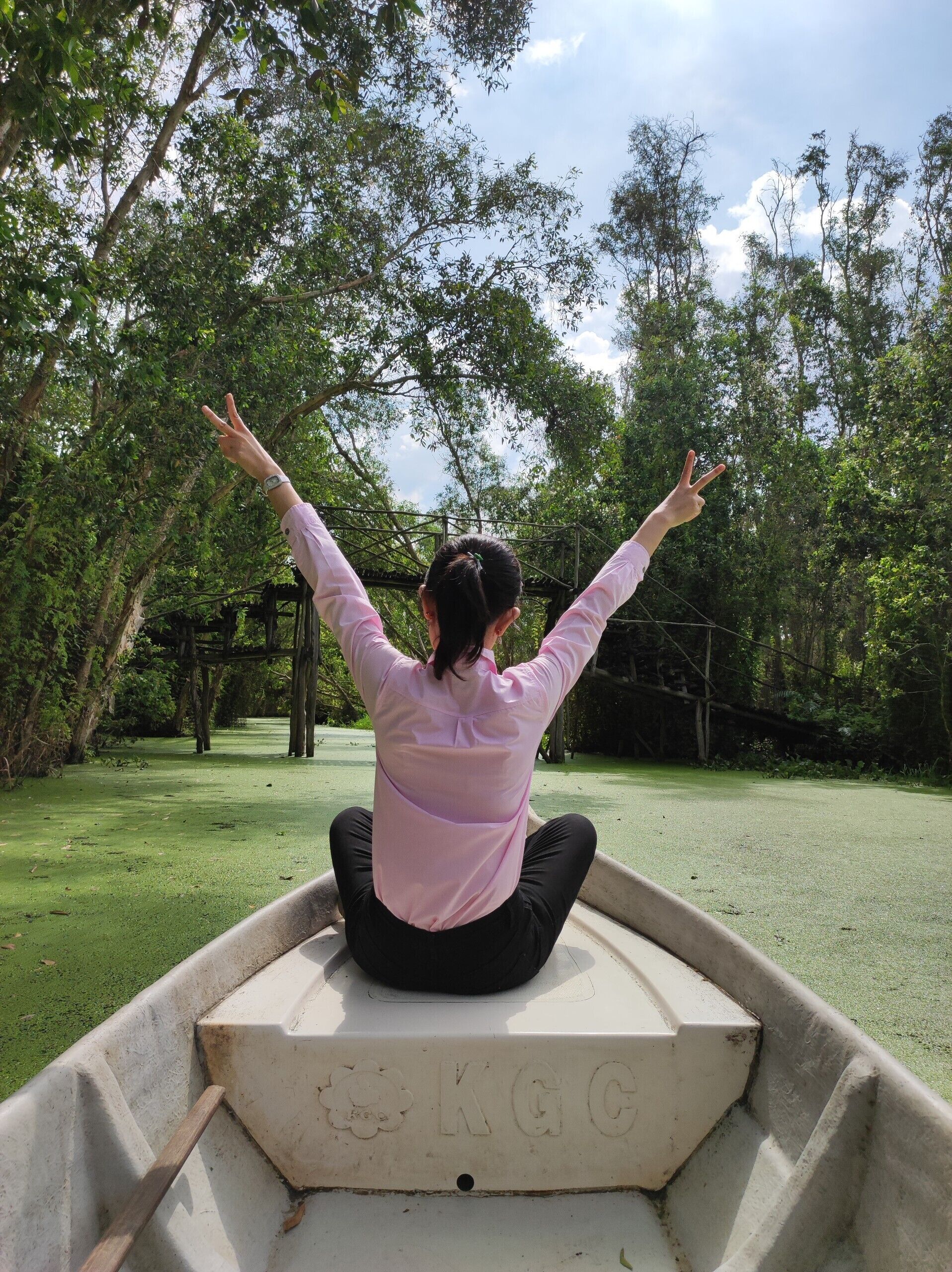 A person sitting in a boat with arms raised, surrounded by greenery and trees.