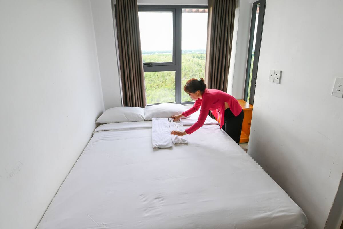Ein Bett mit weißer Bettwäsche, ein Fenster mit Vorhängen und Blick auf Grün.
