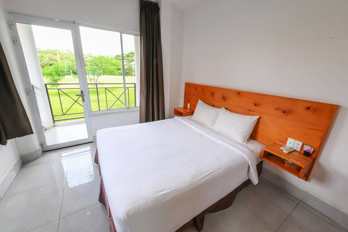 Ein Hotelzimmer mit einem großen Bett, einem hölzernen Kopfteil, einem Nachttisch mit Telefon und Blick auf einen grünen Garten durch eine Schiebetür.