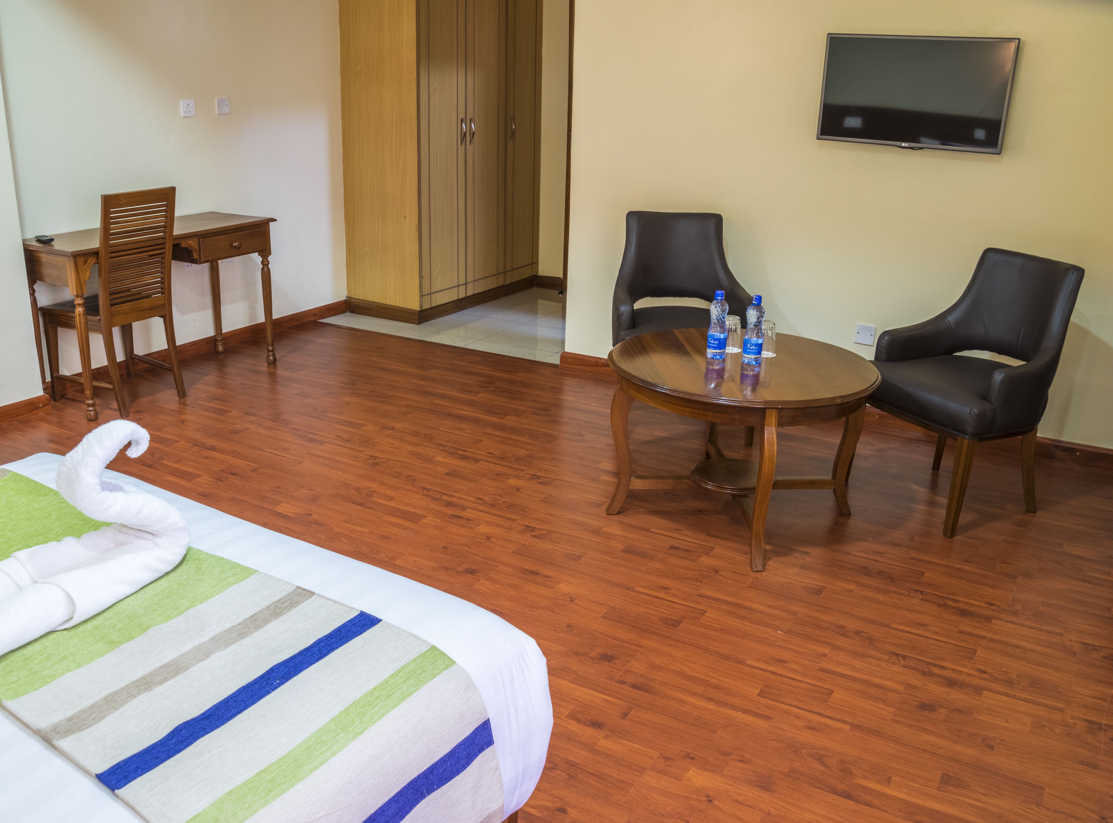 A hotel room with a bed, a wooden desk, two chairs, a small table with water bottles, and a TV on the wall.