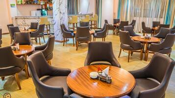 Une salle à manger d’hôtel avec des tables rondes et des fauteuils en cuir, un bar orné d’une colonne en marbre, et une vue sur l’espace bar à travers des rideaux.