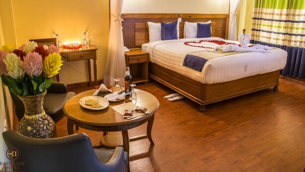 A hotel room with a bed, bedside table, lamp, vase with flowers, and a dining table with a plate and glasses. - Eastmark Hotel (Nakuru)