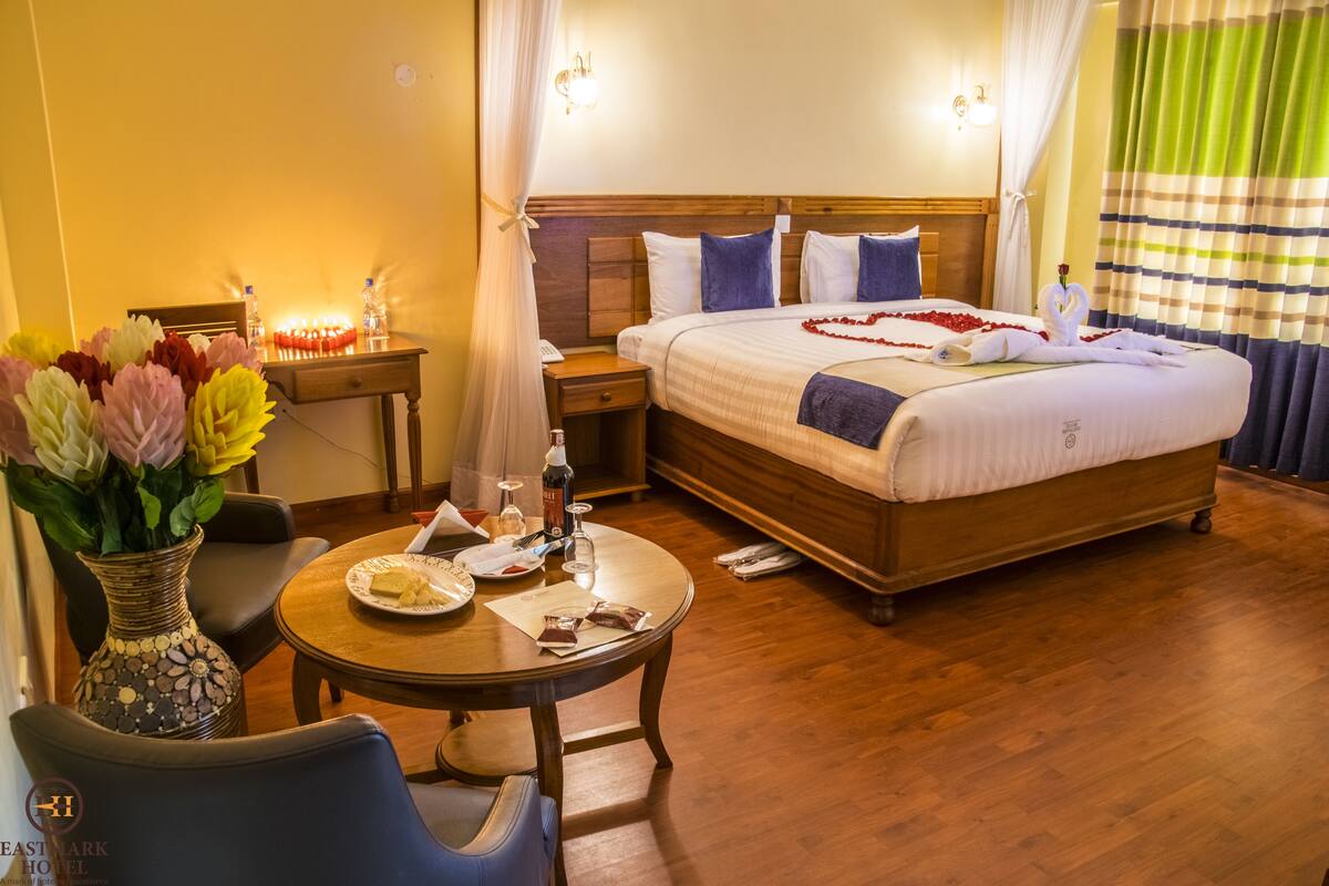 Habitación de hotel con cama, mesita de noche, lámpara, jarrón con flores y una mesa de comedor con plato y vasos.