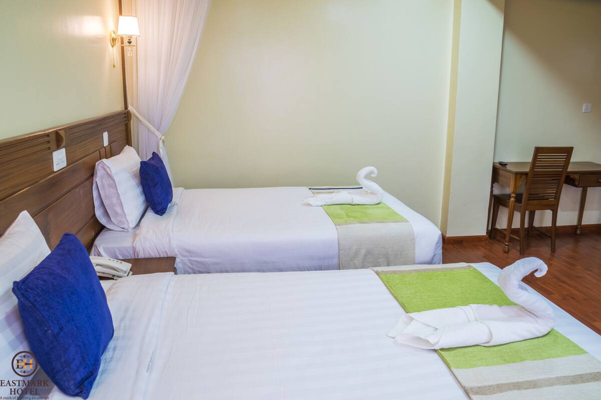Dos camas individuales con sábanas blancas, un cabecero de madera, una toalla verde y blanca, y una silla de madera en una habitación de hotel.