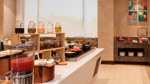 Zona de desayuno tipo buffet con diversas opciones de comida y bebidas, una máquina de café y una ventana con vistas.