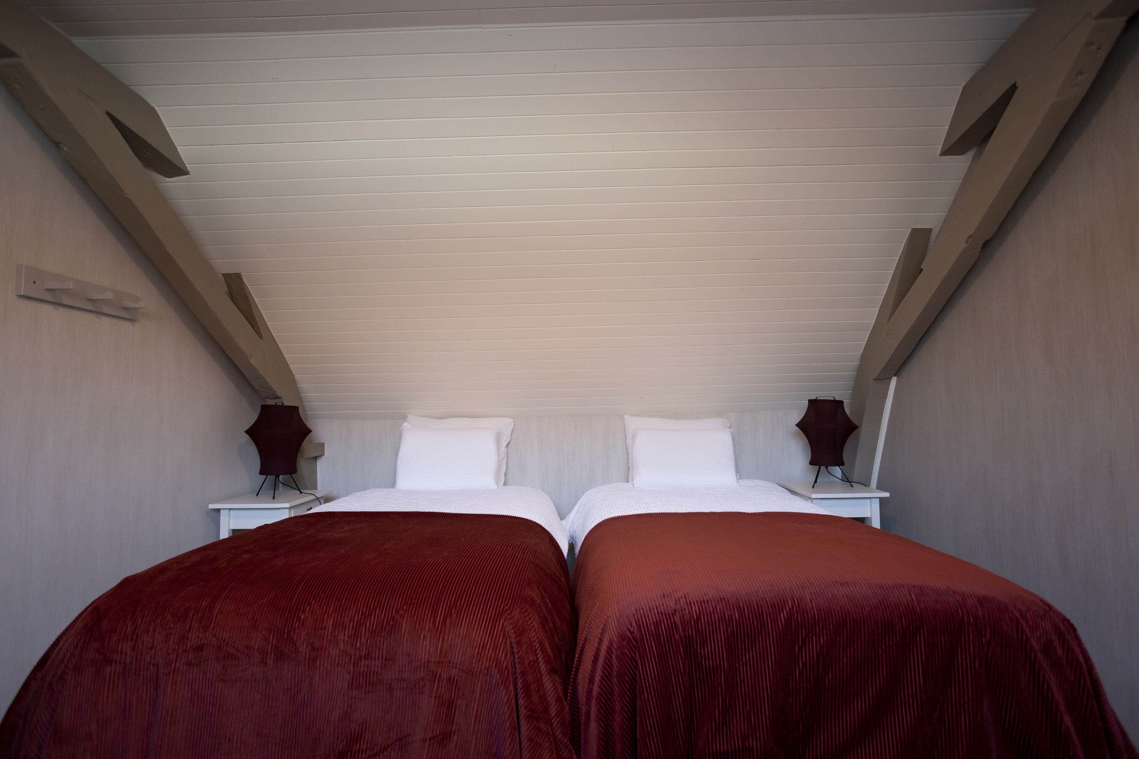 A twin room with two beds, each with a red bedspread, white pillows, and a white headboard. There are two bedside tables with lamps, and the room has a sloped ceiling with wooden beams.